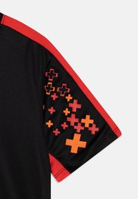 Svart t-shirt med kort ärm och röd trim samt orange och röda plusteckenmönster på ärmen mot en vit bakgrund.