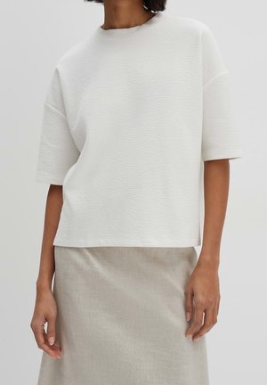 Frau trägt ein strukturiertes weißes Kurzarmoberteil kombiniert mit einem beigen Rock, abgebildet von den Schultern bis zur Mitte des Oberschenkels vor einem einfarbigen Hintergrund.