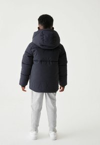 Doudoune marine avec capuche et logo "BAKER" en relief dans le dos, associée à un pantalon de jogging gris et des baskets blanches.