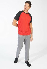 Camiseta atlética de manga corta roja y negra con textura acanalada, combinada con joggers ajustados gris claro que tienen cremalleras y bolsillos laterales, usados con zapatos negros.