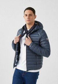 Gevoerde marineblauwe jas met capuchon, voorzien van een ritssluiting, elastische manchetten en een contrasterende, gestreepte voering. Gedragen over een wit shirt.