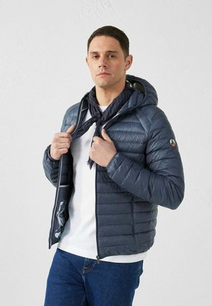 Gevoerde marineblauwe jas met capuchon, voorzien van een ritssluiting, elastische manchetten en een contrasterende, gestreepte voering. Gedragen over een wit shirt.