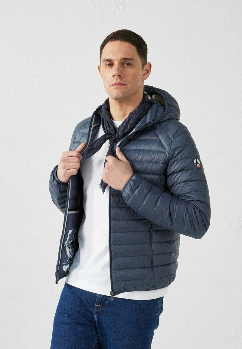 Gevoerde marineblauwe jas met capuchon, voorzien van een ritssluiting, elastische manchetten en een contrasterende, gestreepte voering. Gedragen over een wit shirt.