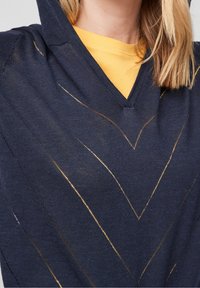 Marinblå hoodie med V-ringning och gyllene metalliska zigzag-mönster. Mjuk textur och lättstickad material som ligger över en gul tröja under.