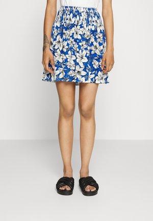 Saia floral azul com padrões de lírios brancos, apresentando uma cintura elástica e bainha ondulada. Combinada com sandálias pretas torcidas.