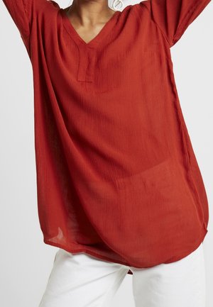 Kvinde iført en løs, tekstureret rød bluse med lange ærmer og V-hals, kombineret med hvide bukser, armene løftet over hovedet.