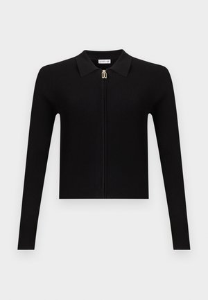 Frau steht in schwarzem langärmligem Reißverschluss-Oberteil und hoch taillierter weiter Hose mit offenen Schuhen vor schlicht weißem Hintergrund.