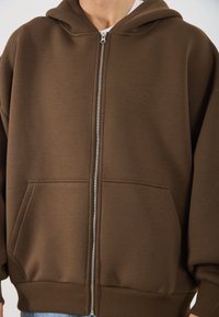 Rjava zip-up kapuca iz mehke tkanine, s sprednjim zadrgo, dvema velikima žepoma in kapuco s vrvico. Riblja manšeta in rob.