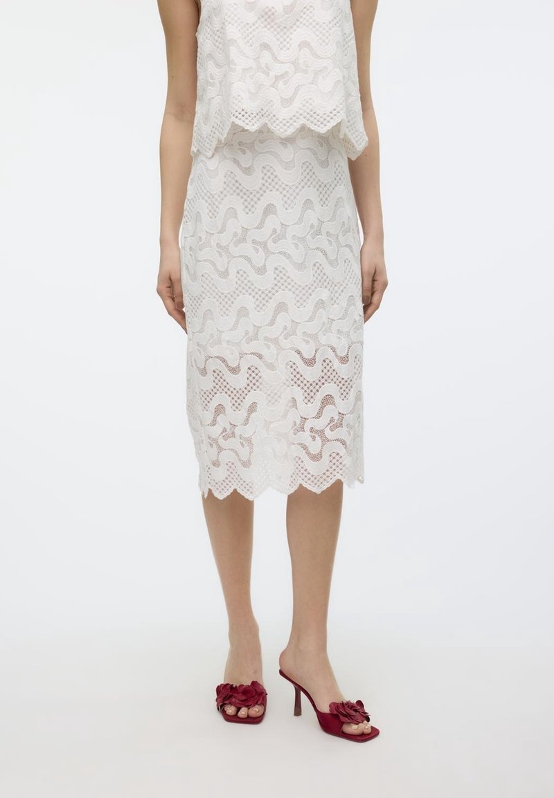 Femme portant une jupe midi en dentelle blanche avec des motifs ondulés et des chaussures ouvertes à talons hauts rouges avec des détails floraux.