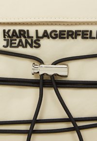 Helle Stoff mit schwarzen elastischen Schnüren, ausgestattet mit einem silbernen Metallverschluss. Schwarze Buchstaben lesen "KARL LAGERFELD JEANS."