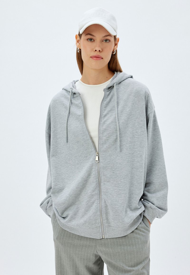 Sudadera con cremallera gris, con capucha y cordones, hecha de material suave; combinada con una camiseta blanca y pantalones grises de rayas.