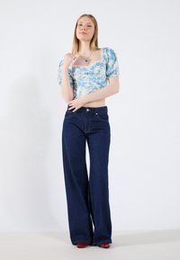 Lyseblå blomstercrop-top med puffærmer, parret med mørkeblå udvidede jeans. Modellen har røde hæle og en sølvhalskæde på.