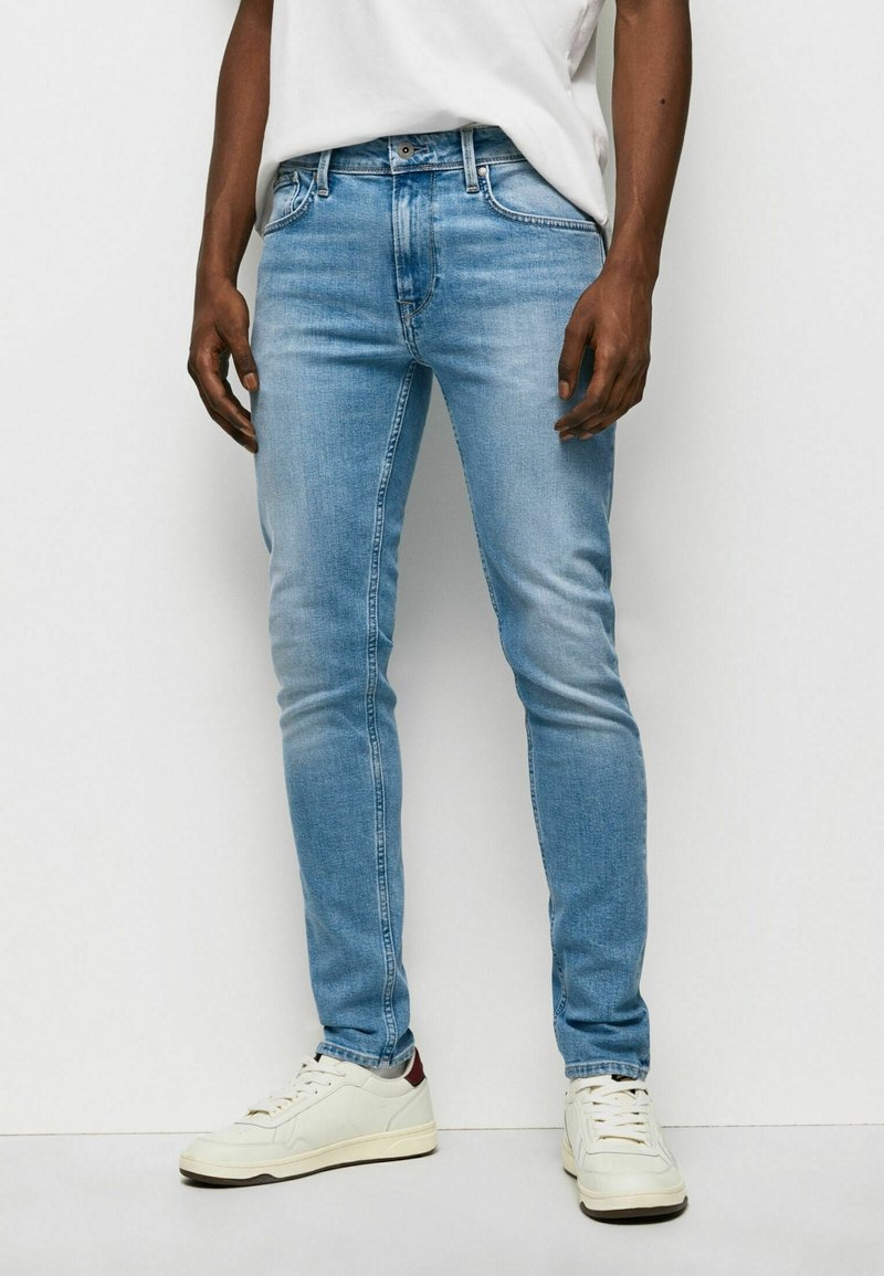 Light blue slim-fit denim jeans with faded patches, five pockets, and a zip fly. Paired with off-white sneakers and a white top.