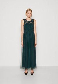 Jauna moteris dėvi tamsiai žalią nėrinių ir permatomą ilgą suknelę be rankovių bei juodus platforminius sandalų, stovinti priešais baltą foną.