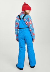 Salopettes de ski bleu rembourrées avec des bretelles réglables, portées par-dessus un haut à rayures colorées. Bonnet rouge avec des accents bleus. Détails de couture visibles.