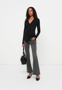 Top nero a maniche lunghe con scollo a V, abbinato a pantaloni grigi a palazzo. La modella tiene in mano una borsa nera e indossa delle scarpe con punta a stiletto nere.