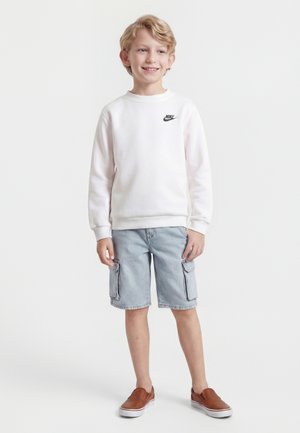 Usmievajúce sa dieťa stojí oblečené v bielej mikine Nike, svetlo-modrých rifľových cargo kraťasoch a hnedých slip-on topánkach na jednoduchom pozadí.