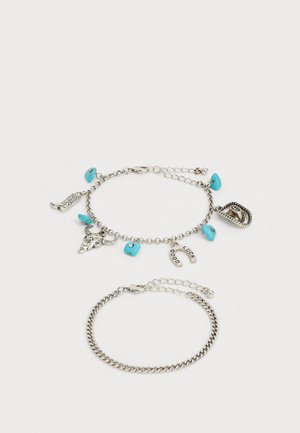 Srebrne narukvice od lanca s tirkiznim perlicama i privjescima, uključujući lubanju i oznake koje kažu "DOBRO" i "SREĆA."