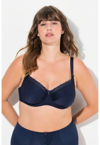 Tummansininen rintaliivi, jossa on pitsireunus, pilkullinen pinta, muotoileva tukilanka ja säädettävät olkaimet. Keskiosassa on koristeellinen nappi.