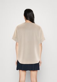 Nainen, jolla on pitkät tummat hiukset, pukeutuneena beigeen lyhythihaiseen löysään puseroon ja mustiin hameeseen, näkyy selkäpuolelta yksinkertaista vaaleaa taustaa vasten.