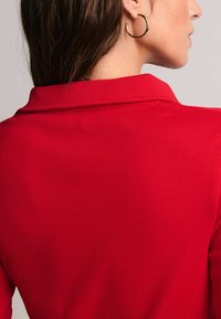 Rød ribbet top med krave, der har en glat tekstur og struktureret design. Synlig guld hoops ørering komplementerer outfittet.