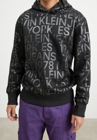 Black hoodie with a textured pattern of white and gray text, featuring "Calvin Klein Jeans" and "New York." Ribbed cuffs and hem.