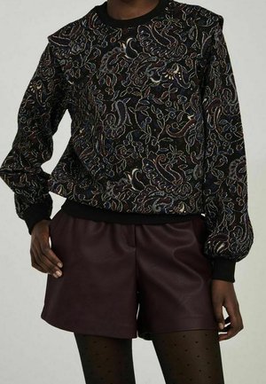 Personne portant un haut à manches longues noir à motif floral, un short en cuir bordeaux foncé, et des collants noirs à petits pois.