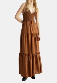 Robe maxi marron avec un corsage en tricot et un ourlet froncé à volants. Elle dispose d'un décolleté en V et de bretelles ajustables, fabriquée dans un tissu doux. Chaussures noires.