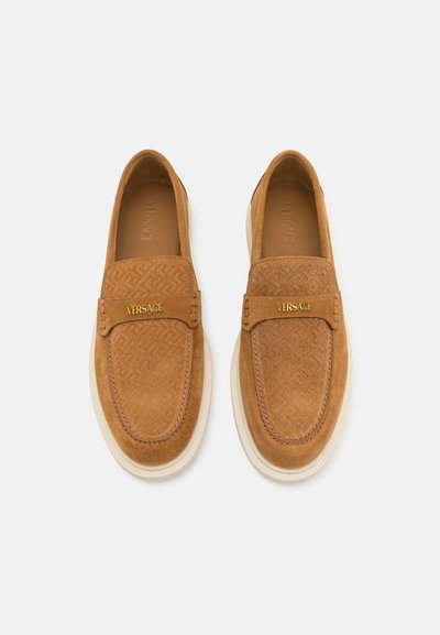 Tan suede slip-on shoes with embossed pattern, featuring a gold logo band and a white rubber sole. Smooth texture throughout.