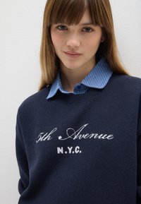Marineblauer Pullover mit weißem Text "5th Avenue N.Y.C." auf der Vorderseite, über einem hellblau gestreiften Hemd getragen. Glatte Textur und lässiges Design.