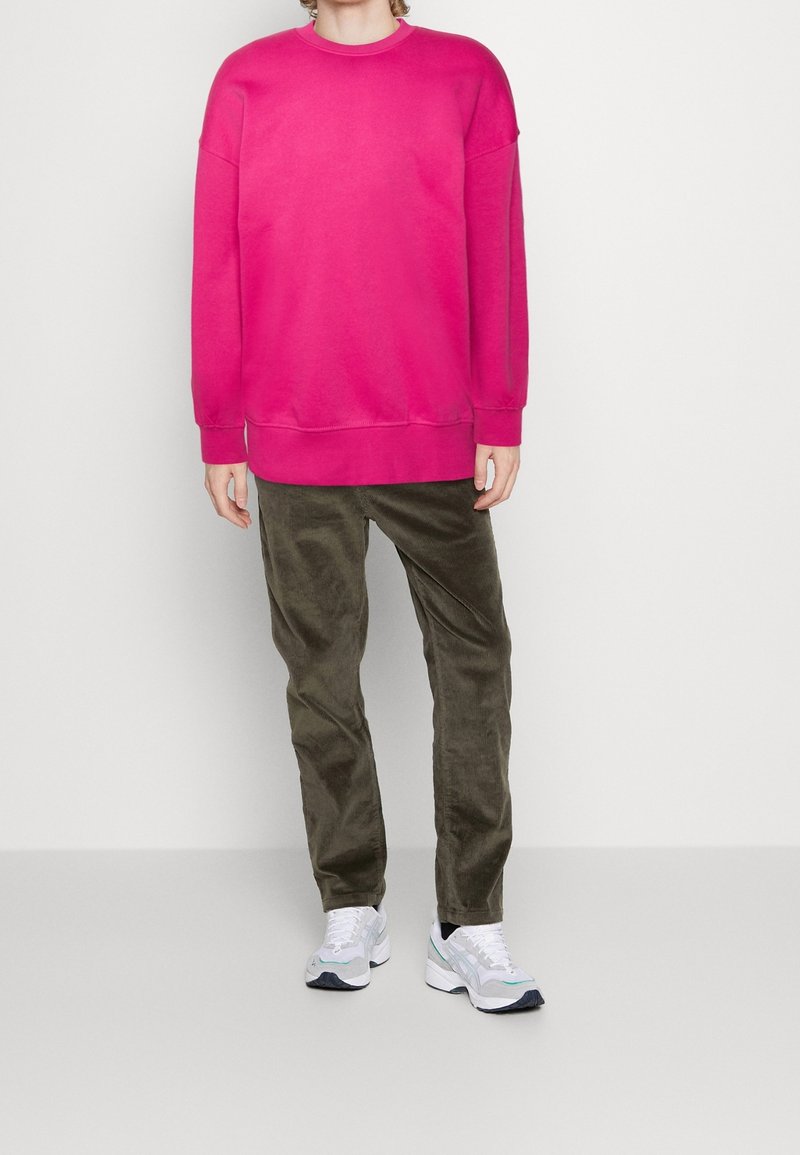 Personne portant un sweat-shirt à manches longues rose vif, un pantalon en velours côtelé olive foncé et des chaussures de sport blanches, debout devant un fond uni.