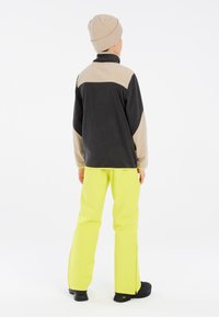 Fleecejack met zwart en beige, gele broek met ritsen, gedragen met een beige gebreide beanie en zwarte laarzen, gezien van achteren.