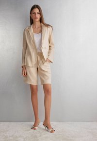 Beige lněný blazer a odpovídající šortky, texturovaná látka, uvolněný střih, doplněné bílými otevřenými muly s černými akcenty.