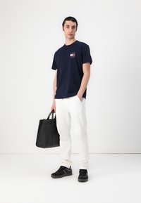 Marineblaues Baumwoll-T-Shirt mit kleinem Logo, kombiniert mit weißen Hosen und schwarzen Sneakern. Hält eine schwarze Ledertasche.