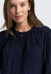 Blouse marine à encolure ronde, avec des détails plissés et une fermeture à bouton en forme de clé à l'avant. Texture de tissu douce avec une silhouette fluide.