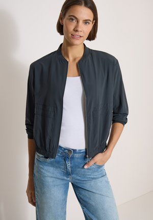 Vrouw met kort bruin haar die een donker jasje met rits draagt over een wit topje en lichtblauwe spijkerbroek, staand met één hand in haar zak.