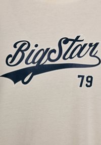Bēša kokvilnas T-kreklu ar tumši zilu "Big Star" tekstu izliektā stilā un skaitli "79" blokveidā. Gluda tekstūra.
