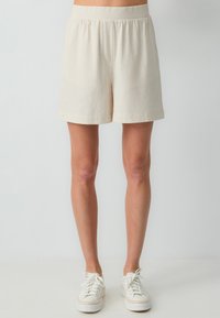 Beige shorts met een elastische tailleband, met een relaxte pasvorm, een zachte textuur en een zoom die boven de knie eindigt. Gepaard met witte sneakers.