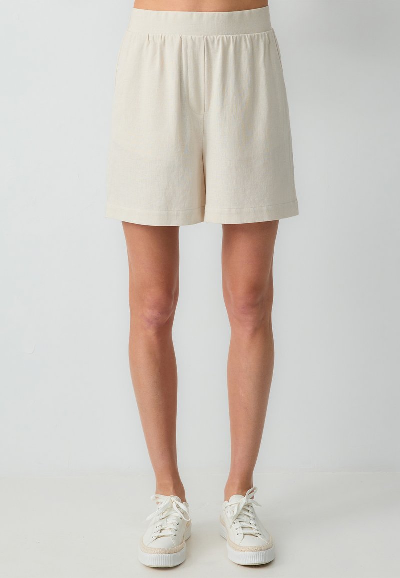 Beige shorts met een elastische tailleband, met een relaxte pasvorm, een zachte textuur en een zoom die boven de knie eindigt. Gepaard met witte sneakers.
