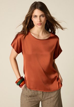 Jonge vrouw met lang bruin haar draagt een losse roestkleurige satijnen blouse en beige geperforeerde broek, geaccentueerd met grote kleurrijke armbanden.