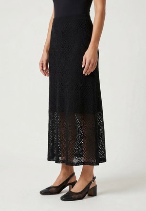 Kobieta w czarnym topie bez rękawów, czarnej koronkowej spódnicy midi z przezroczystą dolną częścią oraz czarnych sandałkach na obcasie z siateczki z paskiem na pięcie i grubym obcasem.