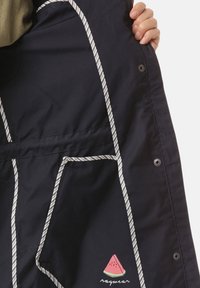 Vnitřek námořnicky modré bundy s bílými pruhovanými akcenty a logem melounu. Má druky a prostornou kapsu. Hladká textilní textura.