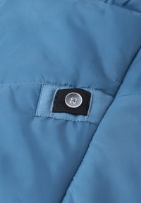 Modrá nadýchaná bunda vyrobená z látky s hladkou textúrou. Obsahuje okrúhly priehľadný gombík na čiernej látkovej nášivke, všitej do materiálu.