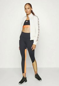 Chaqueta bomber blanca con logo, top corto negro con acentos naranjas y leggings negros con paneles laterales naranja y dorados. Zapatillas deportivas negras.
