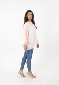 Różowy sweter z szerokimi rękawami i wysokim dekoltem, w połączeniu z dopasowanymi niebieskimi dżinsami i przezroczystymi sandałami na obcasie. Gładka tekstura materiału.
