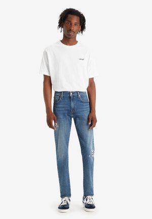 T-shirt bianca in cotone con un piccolo logo nero, abbinata a jeans in denim blu con strappi e taglio dritto. Sneakers blu e bianche.