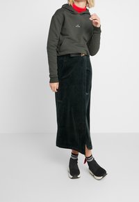 Tmavě zelená mikina s logem ramínka, kombinovaná s sametovou maxi sukní se skluzem po straně a černými nazouvacími botami s pruhovanými ponožkami.