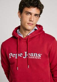 Sudadera de algodón roja con logo, cordones ajustables con acentos plateados y un corte holgado. El fondo es liso.