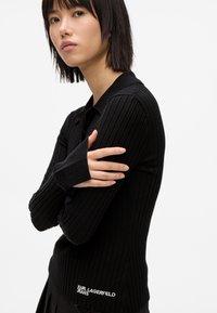 Μαύρο ριγέ πουκάμισο πόλο μακρυμάνικο με γιακά, διαθέτοντας τρύπες για τους αντίχειρες και λεπτομέρεια λογότυπου στο στρίφωμα, κατασκευασμένο από ελαστικό πλεκτό υλικό.