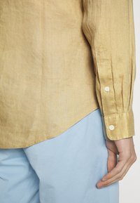 Světle žlutá lněná košile s špičatými manžetami a dvěma knoflíky. V kombinaci se světle modrými kalhotami. Jemná struktura a uvolněný střih.